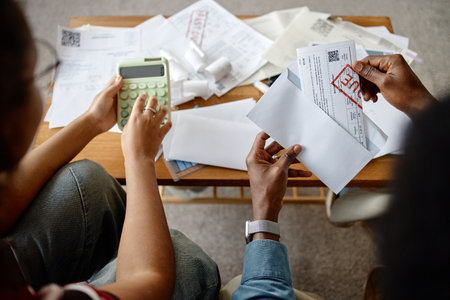 Two adult people, one woman and one Black man, analyzing utility bills and invoices at table, using calculator and opening envelopes, focusing on financial paperworkの写真素材