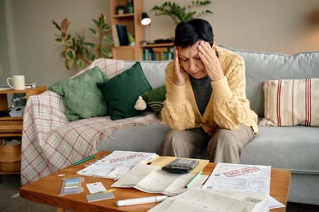 Senior woman sitting on sofa holding head in hands analyzing overdue invoices and bills on table with calculator and documents financial stress on faceの写真素材