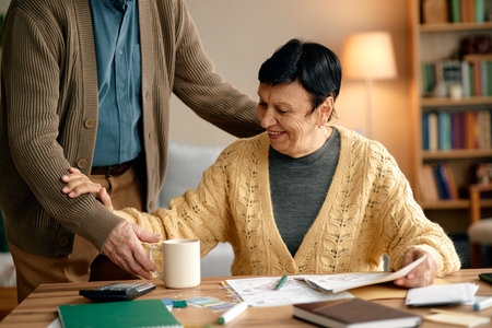 Senior woman smiling while analyzing invoices and bills at table, holding paper in hand, assisted by senior man standing beside her, coffee mug and documentsの写真素材
