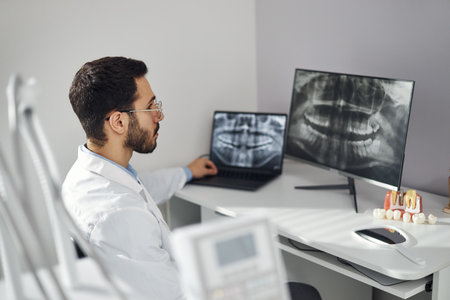 Middle Eastern man wearing lab coat analyzing dental X ray images on computer monitor and laptop in...