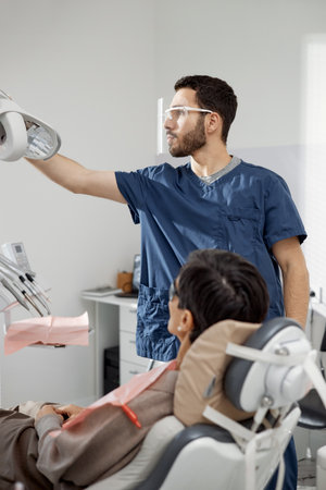Male dentist adjusting dental light while female patient sitting in dental chair, preparing for dental examination in modern clinicの写真素材