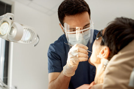Young male dentist examining mouth of middle aged patient during dental appointment, dentist wearing protective mask and gloves, patient reclining in dental chair, clinical setting visibleの写真素材