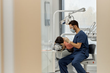 Male dentist treating patient in dental office, dentist wearing medical mask and gloves, patient...