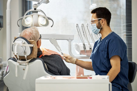 Young male dentist wearing mask and gloves showing mirror to senior Caucasian male patient sitting...