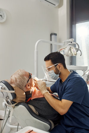 Senior Caucasian man reclining in dental chair, receiving oral examination from young adult male...