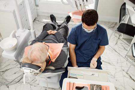 Senior Caucasian man reclining in dental chair receiving treatment from young adult male dentist...