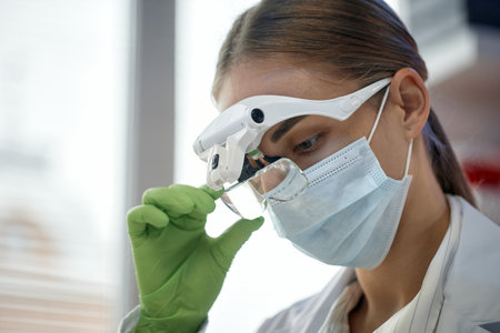 Caucasian young adult woman wearing medical mask and magnifying glasses examining object closely, holding tool with gloved hand, working in laboratory or clinical environmentの写真素材