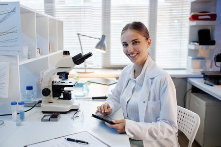 Portrait of young adult Caucasian woman scientist sitting at laboratory desk using digital tablet...