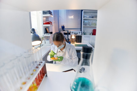 Caucasian young adult woman wearing protective glasses and gloves conducting scientific experiment with petri dish in modern laboratory surrounded by test tubes and equipmentの写真素材