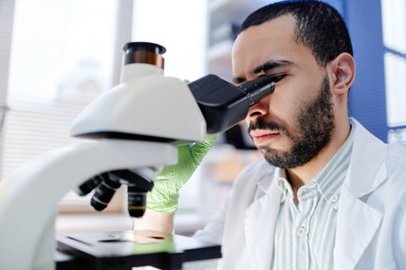 Young adult man examining sample through microscope in laboratory, wearing lab coat and green glove, focusing intently on scientific research in modern workspaceの写真素材