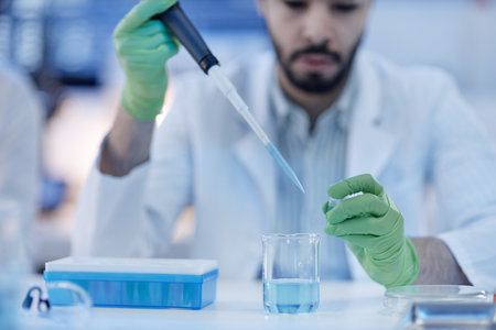 Young adult man wearing laboratory coat and green gloves using pipette to transfer liquid into...