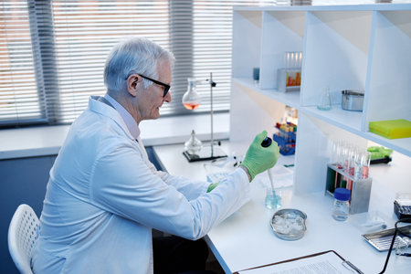 Senior Caucasian man working in laboratory, wearing lab coat and gloves, using pipette to transfer liquid into beaker, surrounded by scientific glassware and equipment on deskの写真素材
