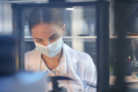 Young adult Caucasian woman wearing medical mask and lab coat working in laboratory, focusing on...