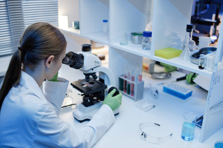 Caucasian young adult woman examining samples using microscope in laboratory setting, wearing lab coat and gloves, surrounded by scientific equipment and glassware on workbenchの写真素材
