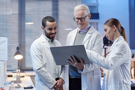 Senior Caucasian man, young adult man, young Caucasian woman wearing lab coats standing together in...