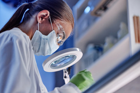 Young adult Caucasian woman wearing protective mask and goggles working in laboratory using magnifying glass, performing scientific research or conducting experimentの写真素材