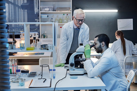Senior Caucasian man supervising young adult man examining sample with microscope in laboratory...