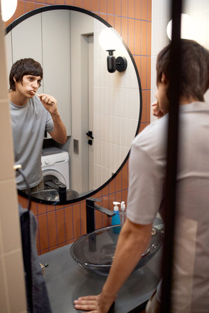 Caucasian man brushing teeth in modern bathroom, standing in front of round mirror with reflection visible, holding toothbrush and looking at own reflection in mirrorの写真素材