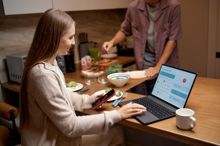 Caucasian young adult woman sitting at kitchen counter using laptop and holding smartphone while man preparing food in background, both engaging in daily home activitiesの写真素材