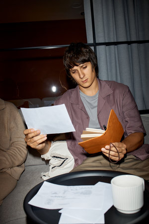Young adult Caucasian man sitting indoors reviewing documents and holding notebook, focused on...