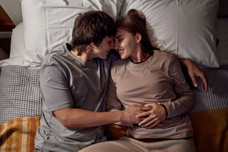 Young Caucasian man embracing young Caucasian woman while lying on bed, both smiling and touching foreheads, couple sharing intimate moment, hands resting on womans stomachの写真素材