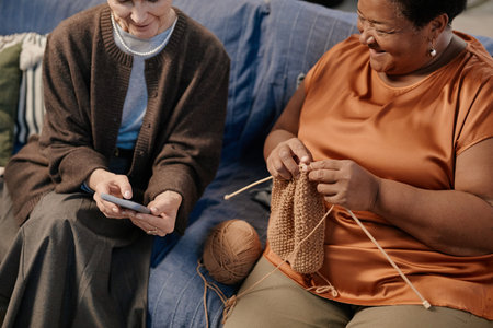 Senior Caucasian woman showing smartphone to senior Black woman knitting yarn, both sitting together...