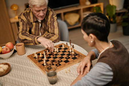Senior Caucasian man playing chess with middle aged woman at table, both concentrating on game,...
