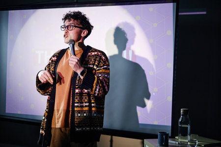 Young adult man standing on stage holding microphone, speaking in front of projected screen during presentation, wearing glasses, curly hair, expressive gestureの写真素材