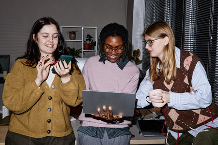 Three young adult people standing together in office, smiling and collaborating while using laptop and smartphone, one holding coffee cup, blinds in backgroundの写真素材