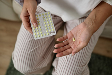 Young adult woman holding blister pack of yellow pills in one hand and two pills in other hand, sitting indoors, showing medication or supplement usageの写真素材
