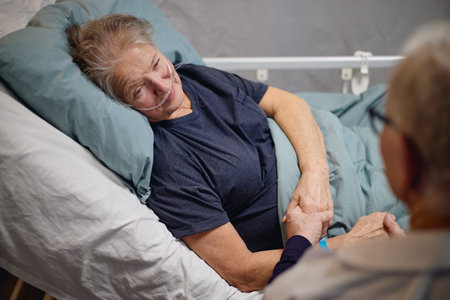 Senior Caucasian woman lying in hospital bed receiving oxygen therapy, holding hands with senior...