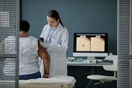 Young adult Caucasian female dermatologist examining upper arm of middle aged Black woman with dermatoscope in medical office, computer monitor displaying skin images in backgroundの写真素材