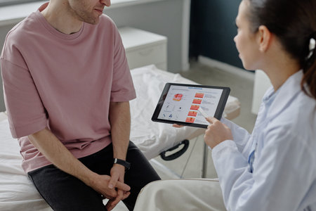 Caucasian young adult man sitting on examination table listening while female doctor showing dermatology information on digital tablet during medical consultation in clinicの写真素材