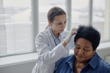 Young adult Caucasian female dermatologist examining scalp of middle aged Black woman in medical office, healthcare professional wearing gloves carefully inspecting patient for dermatological issuesの写真素材