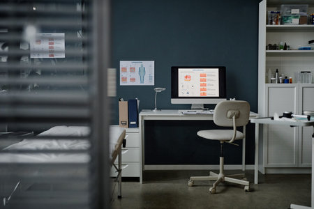 Empty dermatology office showing modern computer displaying skin condition diagrams on desk, examination bed visible in foreground, medical charts and supplies organized on shelves in backgroundの写真素材