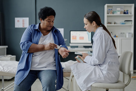 Middle aged Black woman applying moisturizer on skin showing forearm to young adult Caucasian female dermatologist examining and listening patient in medical officeの写真素材