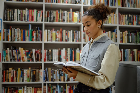 Young adult woman standing in library reading book, focusing on pages while surrounded by shelves...