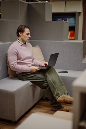 Young adult Caucasian man sitting on sofa using laptop in library, holding smartphone in hand,...