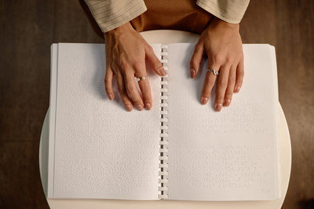 Woman reading braille book with both hands in library, fingers gently moving across raised dots on page, visually impaired accessing information through tactile methodの写真素材