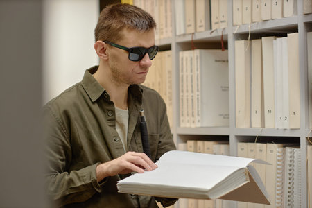 Young adult man wearing sunglasses reading book with hands while standing in library, holding cane,...