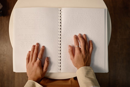 Young adult woman reading braille book with both hands on open pages in library setting, focusing on tactile text, demonstrating accessibility for visually impairedの写真素材