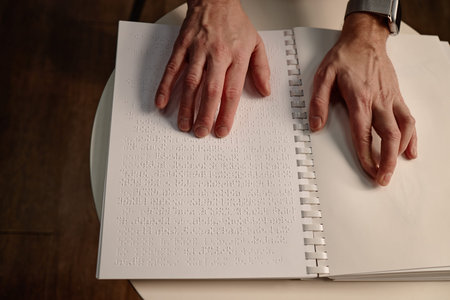 Middle aged man reading braille book with both hands on page, sitting at round table in library,...