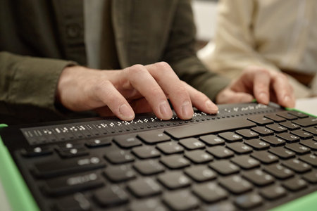 Caucasian young adult man reading braille display with both hands in library setting, focusing on...