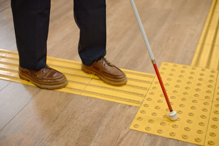 Middle aged Caucasian man using cane for navigation inside library, feet standing on tactile paving...