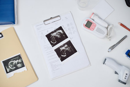 Ultrasound images lying on medical documents with fetal doppler, thermometer, pencil, and folders...