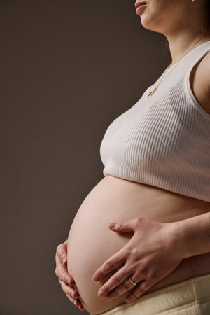 Vertical studio shot of pregnant Caucasian young adult woman holding bare belly with both hands,...