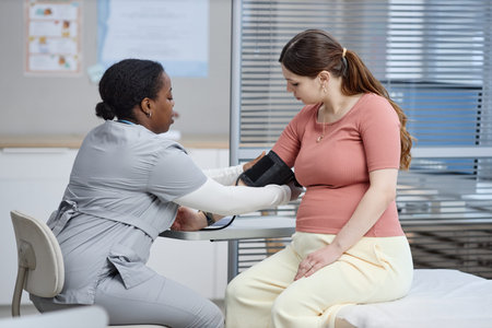 Black woman nurse measuring blood pressure of pregnant Caucasian young adult woman in medical...