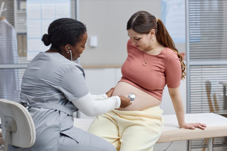 Black female healthcare professional examining pregnant Caucasian young adult woman with stethoscope...