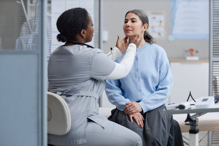 Black woman doctor examining neck of middle aged Caucasian woman during medical checkup in clinic,...
