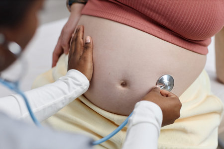 Black female doctor examining pregnant Caucasian woman by placing stethoscope on bare belly,...